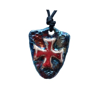 May include: A shield-shaped pendant with a textured, iridescent blue and copper finish, featuring a prominent red cross with white edges in the center. The pendant hangs from a black cord that passes through a hole at the top of the piece.