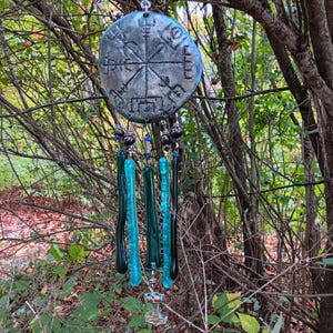 Peut inclure: Carillon artisanal avec un dessus circulaire vieilli orné d'un motif en forme de boussole. En dessous, des brins de tiges de verre turquoise et vert foncé pendent, agrémentés de perles. Un petit pendentif en forme de marteau est suspendu en bas.