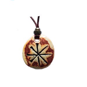 May include: A round, cream-colored ceramic pendant featuring a dark brown, star-shaped etched design with a crackled glaze finish. The pendant is suspended from a dark brown cord and includes a small, decorative bronze-toned metal bead spacer.