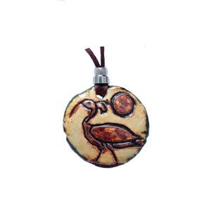 May include: A round pendant necklace with a bird design. The pendant is tan with a dark brown bird and a dark brown circle. The necklace has a brown cord and a silver-colored metal clasp.