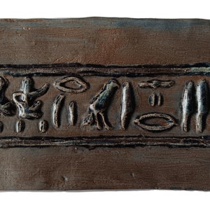May include: A brown clay tile with a raised design of ancient Egyptian hieroglyphs in black and blue. The hieroglyphs are arranged in a horizontal line and include a bird, a circle, and other symbols.