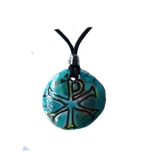 May include: A turquoise pendant with a black cord. The pendant is round with a textured surface and features a black Chi Rho symbol. The pendant is approximately 1.5 inches in diameter.