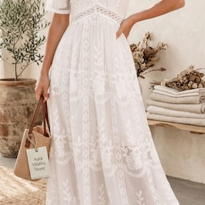 May include: A white floor-length dress featuring short sleeves, a deep V-neckline, and intricate floral lace embroidery throughout. The garment includes a defined waistline with a decorative lace band and a flowing skirt. A small tan woven bag with a tag reading Aura Vitality Store is held at the side.