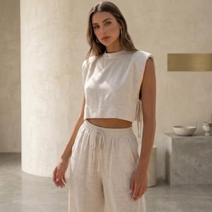 May include: A beige sleeveless crop top with padded shoulders and a high neckline, paired with matching high-waisted wide-leg trousers featuring an elasticated waistband and a drawstring tie. The set is made from a textured, light-colored fabric.
