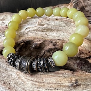 May include: A beaded bracelet featuring pale yellow-green round beads. The bracelet has a central decorative element with an antique silver finish, accented with small, clear crystals. The bracelet is displayed on a textured, light brown wooden surface.