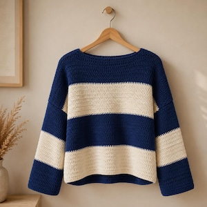 May include: A long-sleeved crochet sweater featuring a horizontal striped pattern in deep navy blue and off-white. The garment has a wide boat neckline, dropped shoulders, and a straight hem, all constructed with a consistent textured stitch.