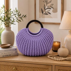 May include: A round, lavender-colored crochet handbag featuring a thick, ribbed texture and two dark brown, circular rigid handles. The bag has a wide, curved silhouette and is constructed from a continuous, dense yarn stitch pattern.