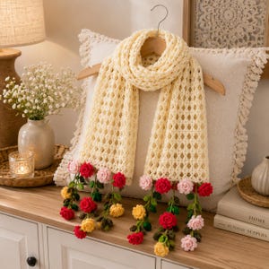 May include: A cream-colored crochet scarf featuring an openwork diamond pattern. The ends of the scarf are decorated with dangling, three-dimensional crocheted flowers in shades of red, yellow, and pink, attached to green crocheted stems.