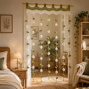 Puede incluir: Una cortina de puerta tejida a ganchillo a mano con un volante superior festoneado en verde oliva y blanco. De la cenefa cuelgan varias tiras verticales, cada una decorada con pequeñas flores de margarita blancas y hojas verdes tejidas.