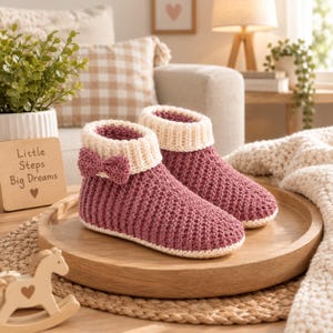 May include: A pair of mauve knitted ankle slippers featuring a textured waffle stitch pattern, a cream-coloured ribbed cuff, and a small matching bow on the side. The slippers are constructed with a solid cream-coloured base and a soft, thick knit fabric.