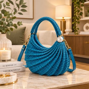 May include: A bright blue crochet handbag with a crescent shape and a textured ribbed pattern. The bag features a single top handle, a detachable blue leather strap with gold-tone hardware, and a large white spherical pearl accent on one side.