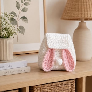 Può includere: Uno zaino in uncinetto bianco con chiusura a patta, due lunghe orecchie da coniglio rosa e bianche sul davanti e una piccola coda a pon-pon bianca centrata tra le orecchie. La borsa è realizzata con un motivo a maglia testurizzato.