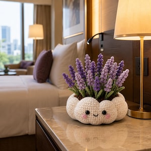 May include: A crocheted lavender plant in a white, cloud-shaped pot with a smiling face. The pot features black bead eyes, a small stitched mouth, and pink blush circles on the cheeks. The lavender stems are shades of purple and green.