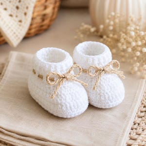 Puede incluir: Un par de patucos de bebé tejidos a ganchillo en color blanco con un patrón de punto texturizado y un lazo de cuerda de yute color canela atado alrededor del tobillo. Los patucos tienen una puntera redondeada y un diseño de caña alta.