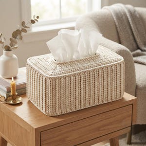 May include: A rectangular tissue box cover crafted from beige yarn in a ribbed crochet pattern. The cover features a central rectangular opening at the top, through which several white paper tissues are pulled upward in a folded arrangement.