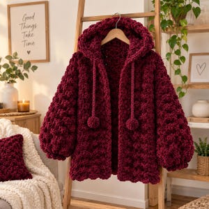May include: A deep burgundy, chunky knit hooded cardigan featuring a textured bobble stitch pattern, long balloon sleeves, and two long drawstrings finished with matching pom-poms at the ends, all displayed on a wooden clothes hanger.