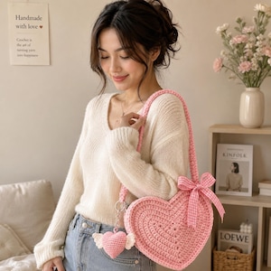 Könnte beinhalten: Eine hellrosa, herzförmige Häkel-Umhängetasche mit strukturiertem Strickmuster, einer passenden rosa karierten Schleife am Träger und einem kleinen herzförmigen Häkelanhänger mit weißen Flügeln an der Seite der Tasche.