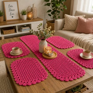 May include: A set of hot pink crocheted table linens consisting of one long rectangular runner and four matching oval placemats. Each piece features a scalloped edge design. Two white ceramic mugs sit on two of the placemats on the wooden table.