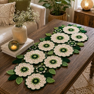Può includere: Un runner da tavola all'uncinetto fatto a mano con un motivo floreale ripetuto, caratterizzato da petali color crema e centri verde scuro, arricchito da piccole foglie all'uncinetto verde scuro tra i motivi floreali più grandi.