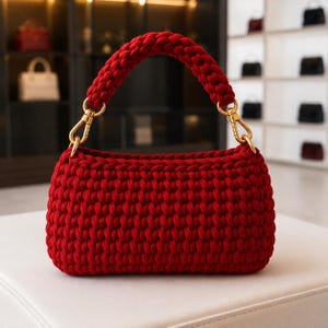 May include: A deep red, hand-crocheted handbag featuring a thick, textured knit pattern. The bag has a matching rounded handle and is attached to the main body with polished gold-tone metal lobster claw clasps and circular rings on each side.