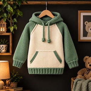 Könnte beinhalten: Ein langärmeliger Strick-Kapuzenpullover mit einem cremefarbenen Mittelteil sowie salbeigrünen Ärmeln, Kapuze und Saum. Das Kleidungsstück verfügt über eine Kängurutasche, gerippte Bündchen und einen passenden Kordelzug mit verknoteten Enden.