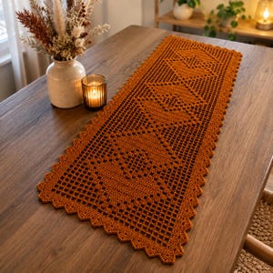 Puede incluir: Un camino de mesa rectangular de ganchillo en color naranja quemado con un patrón geométrico repetitivo de rombos y un borde festoneado. El camino está tejido con hilo fino, creando un diseño de rejilla calada sobre la madera.