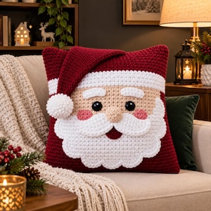 Può includere: Un cuscino quadrato all'uncinetto con il disegno di un volto di Babbo Natale. Il cuscino è principalmente rosso scuro con un bordo bianco e un pompon bianco sull'angolo. Include un naso beige, occhi neri, guance rosa e barba bianca.