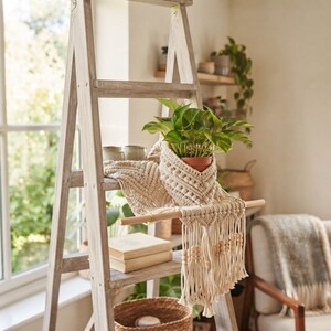 Op de afbeelding: Een crèmekleurige macramé plantenhanger met ingewikkelde geweven patronen en houten kralen, gedrapeerd over een lichte houten ladder. Het macramé-stuk bevat een kleine groene potplant en is afgewerkt met lange franjes aan de onderzijde.
