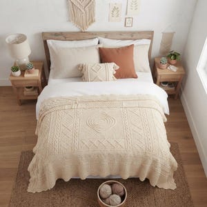May include: A cream-colored knitted throw blanket with intricate cable and diamond patterns draped over a bed. The blanket features a textured surface with a central diamond motif and a scalloped edge, providing a decorative layer for bedding.