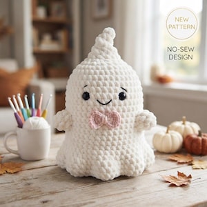 Könnte beinhalten: Ein weißes gehäkeltes Geister-Plüschtier mit schwarzen Sicherheitsaugen, einem gestickten Lächeln und einer kleinen rosa Fliege. Der Text NEW PATTERN NO-SEW DESIGN ist oben rechts sichtbar. Der Geist hat eine texturierte, weiche Garnoptik.