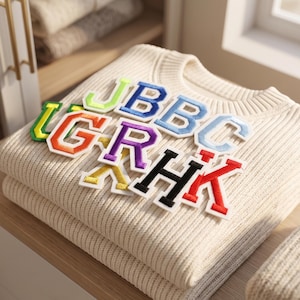 May include: A folded cream-colored ribbed knit sweater featuring a collection of colorful embroidered varsity-style letter patches. The patches include the letters J, B, B, C, L, G, R, H, K, and X in various bright colors like lime, blue, orange, purple, black, red, and yellow.