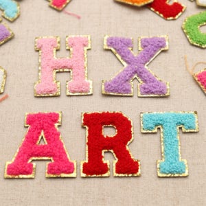 Puede incluir: Una colección de parches de letras individuales con una superficie texturizada y difusa en varios colores, incluyendo rosa, morado, rojo y azul claro. Cada letra está delineada con un borde dorado metálico de estilo bloque.