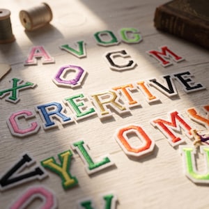 May include: A collection of embroidered fabric letter patches in various colors including pink, green, purple, black, orange, yellow, and blue. The letters are arranged to spell the word CRERTIVE, surrounded by individual letters on a light surface.