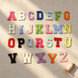Peut inclure: Une collection de vingt-six écussons représentant les lettres de l'alphabet, chacun doté d'une surface texturée et duveteuse dans des couleurs vives comme le rose, blanc, violet, bleu, vert, orange, jaune, rouge et noir, avec un contour doré.
