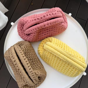 May include: Three rectangular crochet pouches with a thick, textured stitch pattern and integrated handles. One pouch is dusty pink, one is light brown, and one is bright yellow. Each pouch features a structured, hinged-style opening at the top.