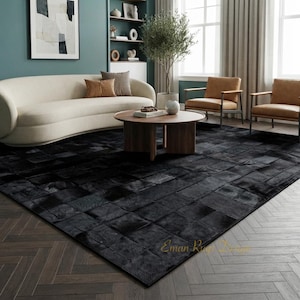 Puede incluir: Una alfombra rectangular grande con un diseño de patchwork compuesto por baldosas cuadradas de cuero negro. La alfombra tiene una textura suave y uniforme y cubre una parte importante del suelo. El texto Eman Rugs Design es visible en oro.