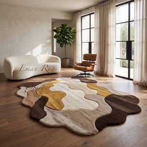 May include: An irregular, organic-shaped area rug featuring a layered, wavy pattern in shades of cream, beige, mustard yellow, and dark brown. The rug has a textured, plush surface and is positioned on a wooden floor. Text reads Eman Rugs Designs.