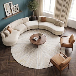 May include: A large, curved, cream-colored sectional sofa with several throw pillows, a round wooden coffee table, and two tan leather armchairs arranged on a circular rug with a radial pattern, set on a dark brown herringbone floor.