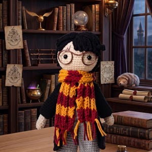 May include: A crochet doll featuring black hair, round brown glasses, and a lightning bolt scar on the forehead. The doll wears a black robe over a grey outfit, a yellow and red striped scarf with fringe, and black and white sneakers.