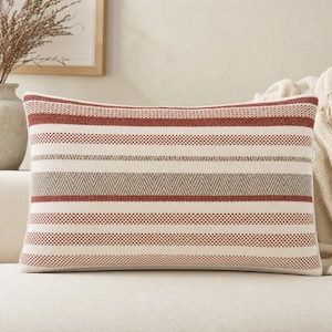 Pode incluir: Uma almofada retangular com um padrão tecido de riscas horizontais em tons de vermelho ferrugem, creme e bege. O design inclui bandas alternadas de cor sólida, pequenas texturas axadrezadas e um padrão central tecido em estilo espinha.