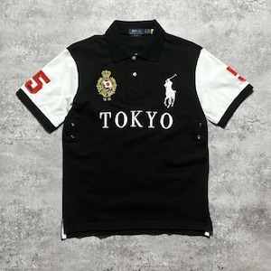 May include: A black polo shirt with white sleeves, featuring the word TOKYO in white letters across the chest. It includes a white embroidered polo player logo, a gold crest with a Japanese flag, and a red number 5 on the right sleeve.