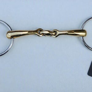 May include: A horse bit featuring a gold-coloured metal mouthpiece with a central joint and two large, silver-coloured circular rings on each end. A small black rectangular tag with the white text GallopGrip is attached to one of the rings.