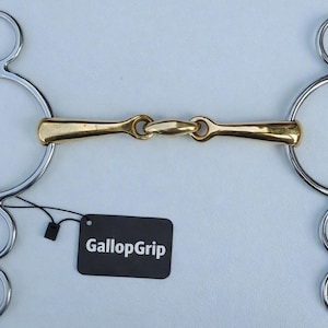May include: A silver-colored metal horse bit featuring a gold-colored mouthpiece with a central joint. The bit has large circular side rings with smaller rings attached above and below. A black rectangular tag with the white text GallopGrip is attached.