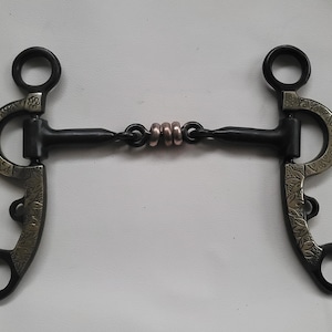 May include: A black metal horse bit featuring two side shanks with intricate silver-toned floral engravings. The mouthpiece consists of two black bars connected in the centre by three small, smooth copper-coloured rings that allow for movement.