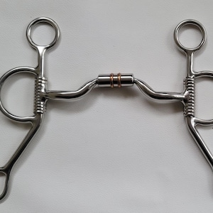May include: A silver-coloured metal horse bit featuring a jointed mouthpiece with two copper-coloured rings in the centre. The bit includes two large side shanks with integrated rings and a series of horizontal grooves along the vertical bars.