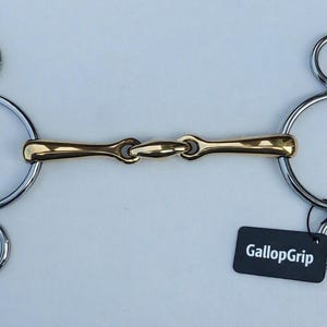 Può includere: Un imboccatura per cavalli con un pezzo di metallo dorato con giunto centrale e guance in metallo argentato con tre anelli su ciascun lato. Un'etichetta rettangolare nera con il testo bianco GallopGrip è attaccata a un anello.