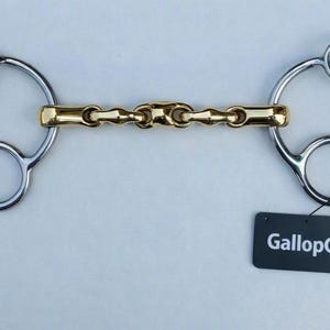 May include: A horse bit featuring a gold-colored metal mouthpiece with a multi-link design connected to two silver-colored metal cheekpieces, each with three rings. A black rectangular tag with the white text GallopGrip is attached to one ring.