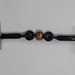 May include: A horse bit featuring two large silver-coloured metal rings connected to a black mouthpiece. The centre of the mouthpiece includes two black links and a single copper-coloured bead, creating a jointed design for equestrian use.