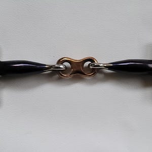 Pode incluir: Um freio para cavalo com dois anéis metálicos prateados em cada extremidade, conectados a barras azul-escuras de superfície lisa que se encontram no centro com um pequeno elo metálico oval de cor cobre, criando um bocal de articulação dupla.