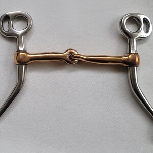 Könnte beinhalten: Ein Pferdegebiss mit einem kupferfarbenen, gelenkigen Mundstück, das mit zwei silberfarbenen Metallschenkeln verbunden ist. Die Schenkel haben einen oberen Ring für das Kopfstück und einen unteren Ring für die Zügel, mit geschwungenem Design.